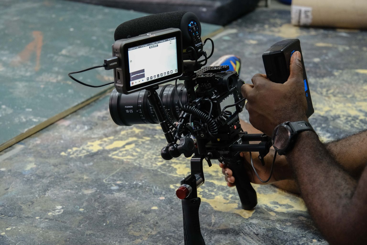 Man's hands holding a professional video camera with a mounted monitor, adjusting settings on a textured floor background. Man's hands holding a professional video camera with a mounted monitor, adjusting settings on a textured floor background.
