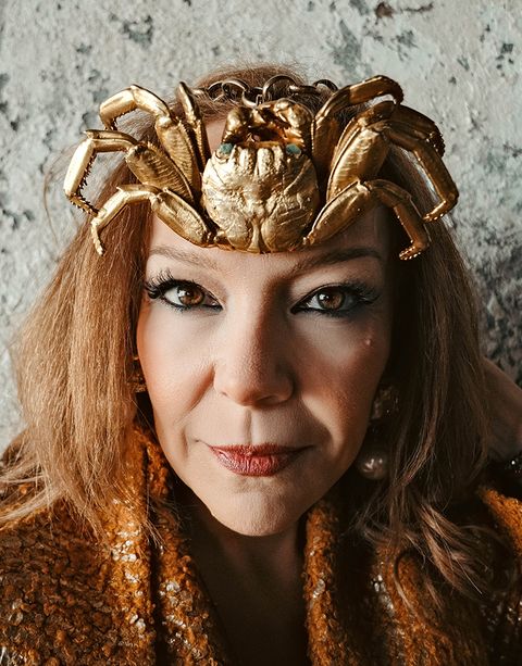Woman wearing a gold crab headpiece and sequined outfit, posing confidently against a textured background.
