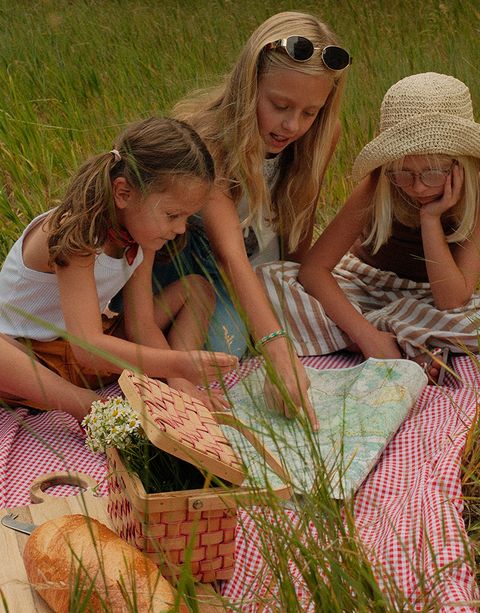 Three children sitting on a picnic blanket in a grassy field, looking at a map. A basket and bread are nearby.