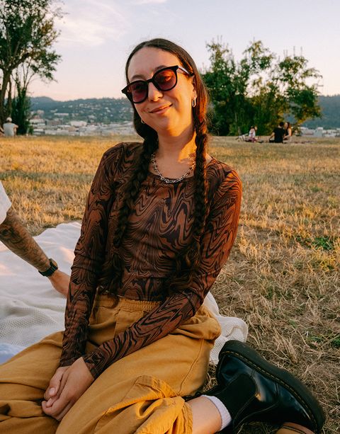 A person with long braided hair and sunglasses sits on a blanket in a grassy field, smiling. Trees and a cityscape are in the background.