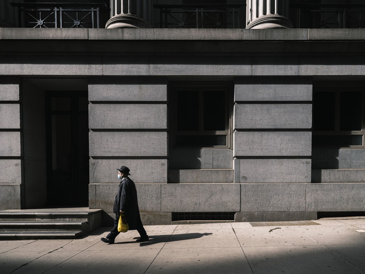 Person in dark coat and hat walks on a sunlit sidewalk beside a large, shadowed stone building, carrying a yellow bag. Person in dark coat and hat walks on a sunlit sidewalk beside a large, shadowed stone building, carrying a yellow bag.